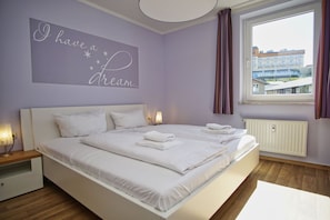 A bedroom with a double bed, a nightstand, a wall-mounted light, a window with curtains, and a view of buildings outside.