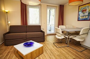 A modern room with a brown sofa, a white table with a blue bowl, a dining table with chairs, a floor lamp, and a painting on the wall.