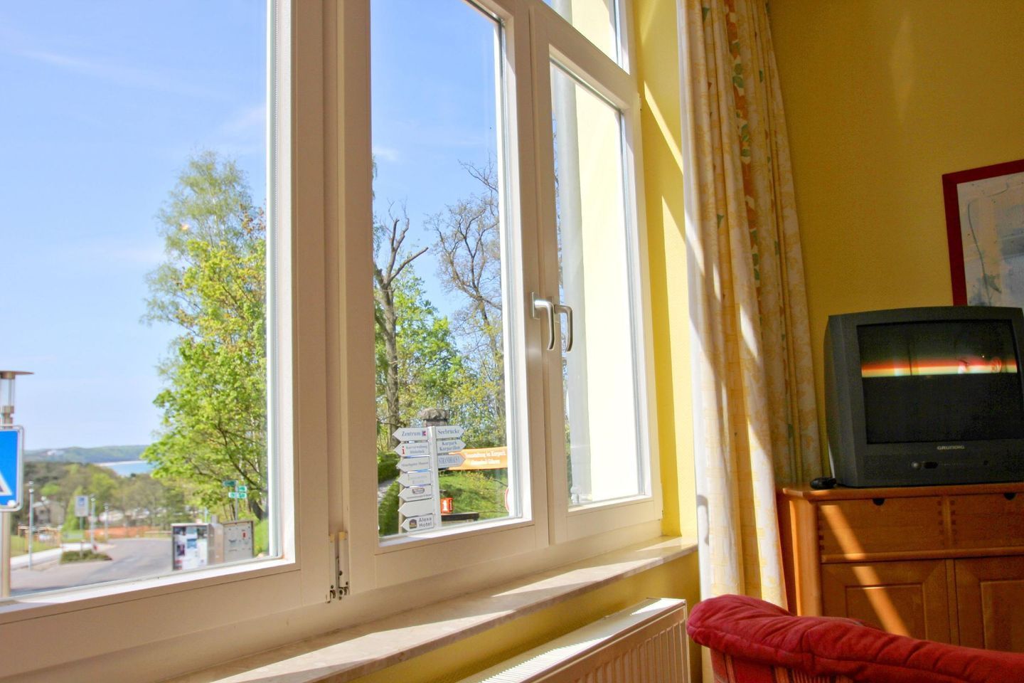 A room with a large window, a view of trees and a road, a television on a wooden stand, and a red chair.