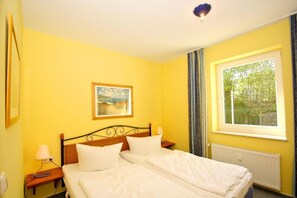 A room with yellow walls, a bed with white bedding, a painting on the wall, and a window with a view of trees. - Close to the beach, free sauna & swimming pool - Strandresidenz Brandenburg FeWo 22 - House (Göhren)
