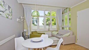 Une pièce moderne avec une table de salle à manger ronde, des chaises blanches, un canapé et une grande fenêtre avec des rideaux.