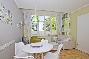 A modern room with a round dining table, white chairs, a sofa, and a large window with curtains. - Close to the beach, balcony, free AHOI adventure pool - Strandresidenz Brandenburg FeWo 24 (Göhren)