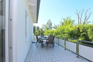 Uma varanda com chão de azulejos, mesa e cadeiras, e vista para árvores verdes e céu claro.