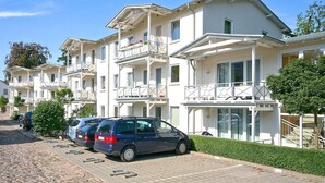 A row of white multi-story buildings with balconies, parked cars, and a paved walkway. - Close to the beach, balcony, free AHOI adventure pool - Strandresidenz Brandenburg FeWo 04 (Göhren)