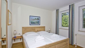 A neatly made bed in a room with a wooden headboard, a bedside table with a lamp, a framed picture on the wall, and a window with curtains. - Close to the beach, balcony, free AHOI adventure pool - Strandresidenz Brandenburg FeWo 04 (Göhren)