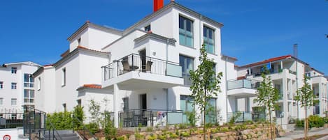 A modern multi-story residential building with balconies, a stone retaining wall, and a clear blue sky.