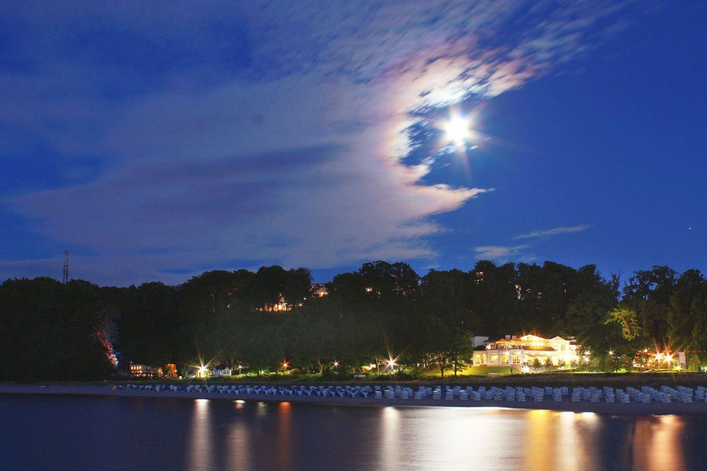 A lakeside view at dusk with a prominent moon and stars in the sky, a calm water surface reflecting lights, and a distant illuminated building surrounded by trees.