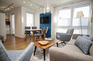 Ein modernes Wohnzimmer mit Essbereich, einer Küche im Hintergrund und großen Fenstern mit Jalousien.