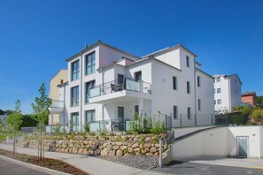 A modern multi-story residential building with balconies, a stone retaining wall, and a paved walkway.