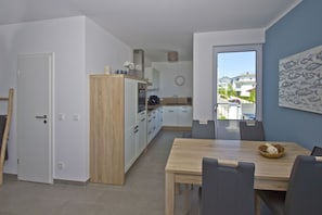 Eine moderne Küche mit weißen Schränken und einer Holzarbeitsplatte, ein Essbereich mit Holztisch und Stühlen sowie Blick auf Häuser durch ein großes Fenster.