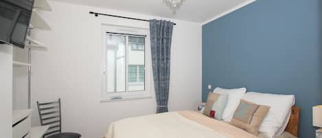 A bedroom with a bed, a chair, a window with curtains, a chandelier, and a bedside table.