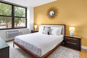 Quarto com uma grande janela, uma cama com roupa de cama branca, duas mesas de cabeceira com abajures e um espelho em formato de sol na parede.