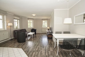 A spacious living area with a sofa, a dining table, and chairs. There are windows with blinds, a radiator, and a lamp.