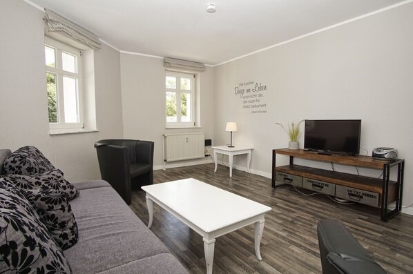 Un salon moderne avec un canapé gris, une table basse blanche, un fauteuil en cuir noir, un téléviseur posé sur un meuble en bois et un radiateur.