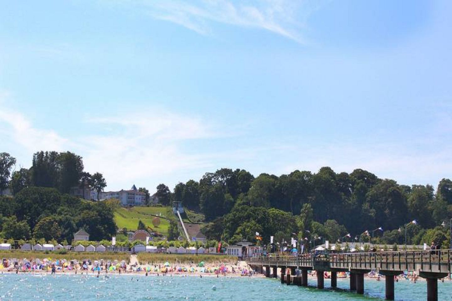 A beach scene with a pier extending into the water, a crowd of people on the beach, and buildings in the background.