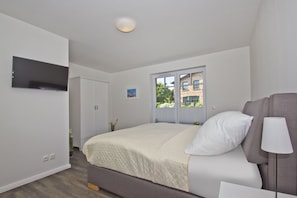 A bedroom with a bed, a TV mounted on the wall, a window with a view of buildings, and a bedside lamp.