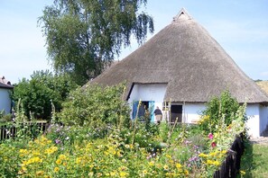 A thatched-roof house with a white exterior, surrounded by a garden with various flowers and a tall tree. - Cozy vacation apartment with terrace and garden - Ferienhaus Zaunkönig FeWo EG (Gager)