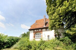 A traditional house with a red-tiled roof, white walls, and arched windows, surrounded by lush greenery.
