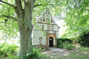Une église à la façade en briques, dotée de fenêtres en arc de cercle et d’un clocher, le tout entouré d’une végétation luxuriante.