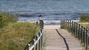 Ein hölzerner Steg führt zu einem Strand, in der Ferne ist eine Person zu sehen.