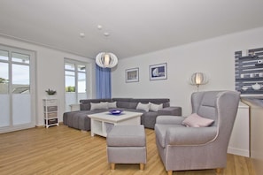 A modern living room with a grey sofa, a matching armchair, a coffee table, and a side table. There is a large window with a view of the outside, a ceiling light, and a wall-mounted shelf with decorative items.