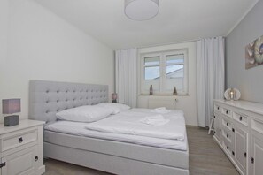 Een netjes ingerichte slaapkamer met een groot bed, een nachtkastje met lamp, een dressoir met lades en een raam met gordijnen.