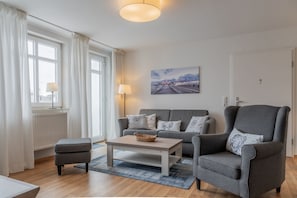 A modern living room with a grey sofa, a matching armchair, a coffee table, and a painting on the wall. - Die Ferienwohnung Nr. 14 im Haus Strandeck - Haus Strandeck (Göhren)