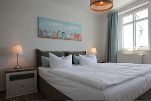 A bedroom with a large bed, two bedside tables with lamps, a painting of beach huts, and a window with curtains. - Die Ferienwohnung Nr. 14 im Haus Strandeck - Haus Strandeck (Göhren)