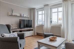 A living room with a flat-screen TV, grey sofas, a coffee table, and a window with curtains. - Die Ferienwohnung Nr. 14 im Haus Strandeck - Haus Strandeck (Göhren)
