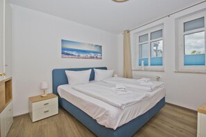 A bedroom with a bed, a nightstand, a lamp, a window with curtains, and a painting on the wall. - Die Ferienwohnung Nr. 07 im Haus Strandeck - Haus Strandeck (Göhren)
