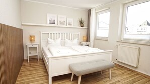 Une chambre à coucher blanche, avec un lit, des tables de chevet, un banc, une fenêtre et un radiateur.