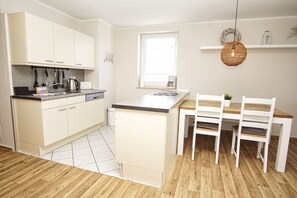 Een keuken met witte kasten, een houten eettafel met stoelen, een hangende lamp en een raam.