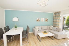A modern living room with a white dining table, grey sofa, and a small coffee table. There are three framed pictures on the wall and a potted plant on the table. - Haus Nordstrand - Vacation Apartment with Terrace, Pool, and Sauna Access - House (Göhren)