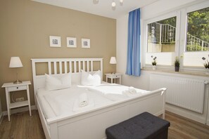 A bedroom with a white bed, two bedside tables, a window with a view of greenery, and a radiator. - Haus Nordstrand - Holiday apartment with terrace, use of swimming pool and sauna - House (Göhren)