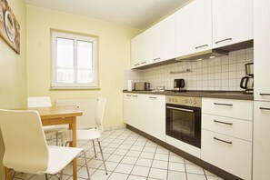 A kitchen with white cabinets, a stove, a microwave, and a coffee maker. There is a window with blinds, a radiator, and a table with chairs. - Haus Rügen - Vacation apartment with a balcony, access to the pool and sauna - Haus Rügen (Göhren)