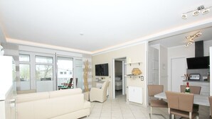 A modern living room with a sofa, a dining table with chairs, a TV mounted on the wall, and a kitchen area with a microwave and toaster. - FeWo Ostseeperle - with sea view and balcony, use of swimming pool and sauna - FeWo (Göhren)