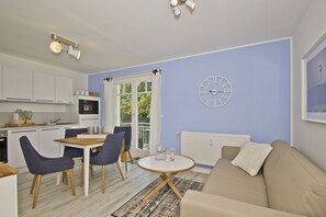 Un salon moderne avec un canapé beige, une table à manger avec des chaises, un coin cuisine avec des meubles blancs, une horloge au mur et une fenêtre avec des rideaux.