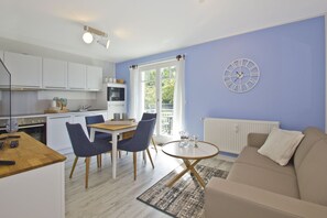 Une cuisine moderne avec des meubles blancs, un coin repas avec une table en bois et des chaises bleues, une table basse ronde et un canapé beige.