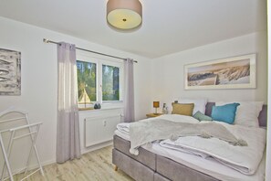 A bedroom with a bed, a radiator, a window with curtains, a nightstand with a lamp, and a painting on the wall.