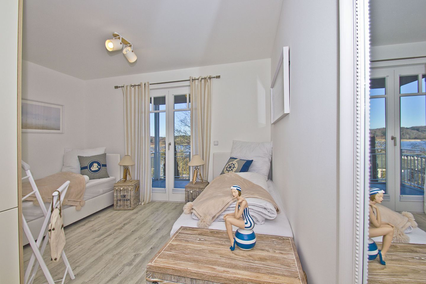 A bedroom with a bed, a chair, a small table, and a dresser. There is a window with curtains and a view of the outside.