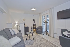 Una sala de estar moderna con un sofá gris, una mesita redonda pequeña, una lámpara de pie y una silla. Hay una estantería y un televisor montado en la pared.