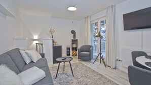 A modern living room with a grey sofa, a small round table, a floor lamp, and a chair. There is a bookshelf and a TV mounted on the wall.