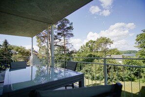 Un balcón con una mesa y sillas de vidrio, con vistas a un lago y una zona boscosa.