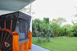A wicker chair with a wooden frame and a cushioned seat, positioned on a concrete patio with a grassy lawn and various plants in the background. - Vacation apartment with a terrace, fireplace, and sauna - Ferienresidenz Zwei Bodden FeWo 2-6 - (Lietzow)