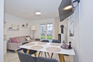 A modern living room with a sofa, a dining table with placemats, a flat-screen TV mounted on the wall, and a large window with a view of the outdoors. - Vacation apartment with a terrace, fireplace, and sauna - Ferienresidenz Zwei Bodden FeWo 2-6 - (Lietzow)