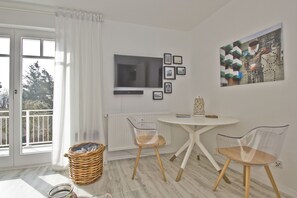 A modern room with a large window, a round table with two chairs, a radiator, and a wall-mounted TV. - Modern 2-room vacation apartment with balcony - Ferienresidenz Zwei Bodden FeWo 1-4 - (Lietzow)