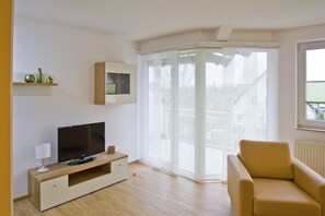 A modern living room with a flat-screen TV on a wooden TV stand, a beige sofa, and a sliding glass door with a floral pattern. - Close to the beach, with a balcony; access to the pool and sauna - Strandhaus Mönchgut FeWo13 (Middelhagen)