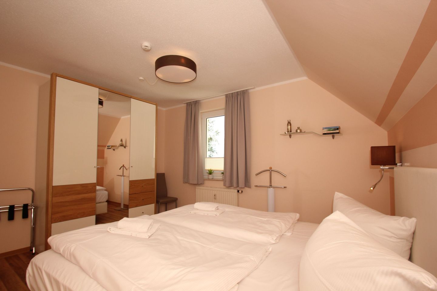 Une chambre d’hôtel avec un grand lit, un miroir, une fenêtre avec des rideaux, une table de chevet et une étagère fixée au mur.