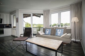 A modern living room with a sofa, armchair, and coffee table. There is a kitchen area with a microwave and cabinets. Large windows provide a view of the outdoors. - Close to the beach with balcony, use of swimming pool and sauna - Strandhaus Mönchgut FeWo11 (Middelhagen)