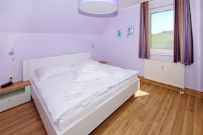 A neatly made bed with white linens and towels, a bedside table with a lamp, and a window with curtains.